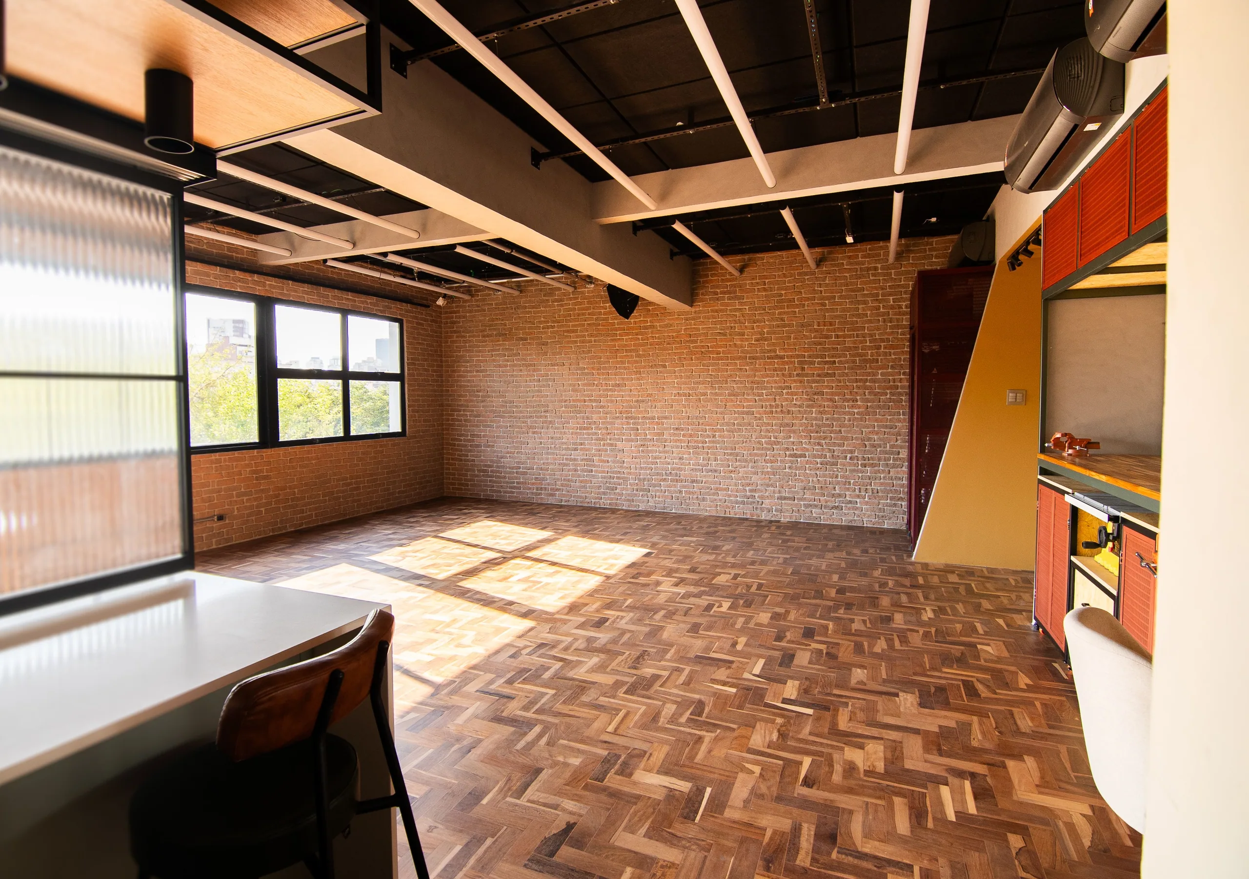 Sala vazia com parede de tijolo e janela para a cidade, bancada de apoio à esquerda e bancada de marcenaria à direita.