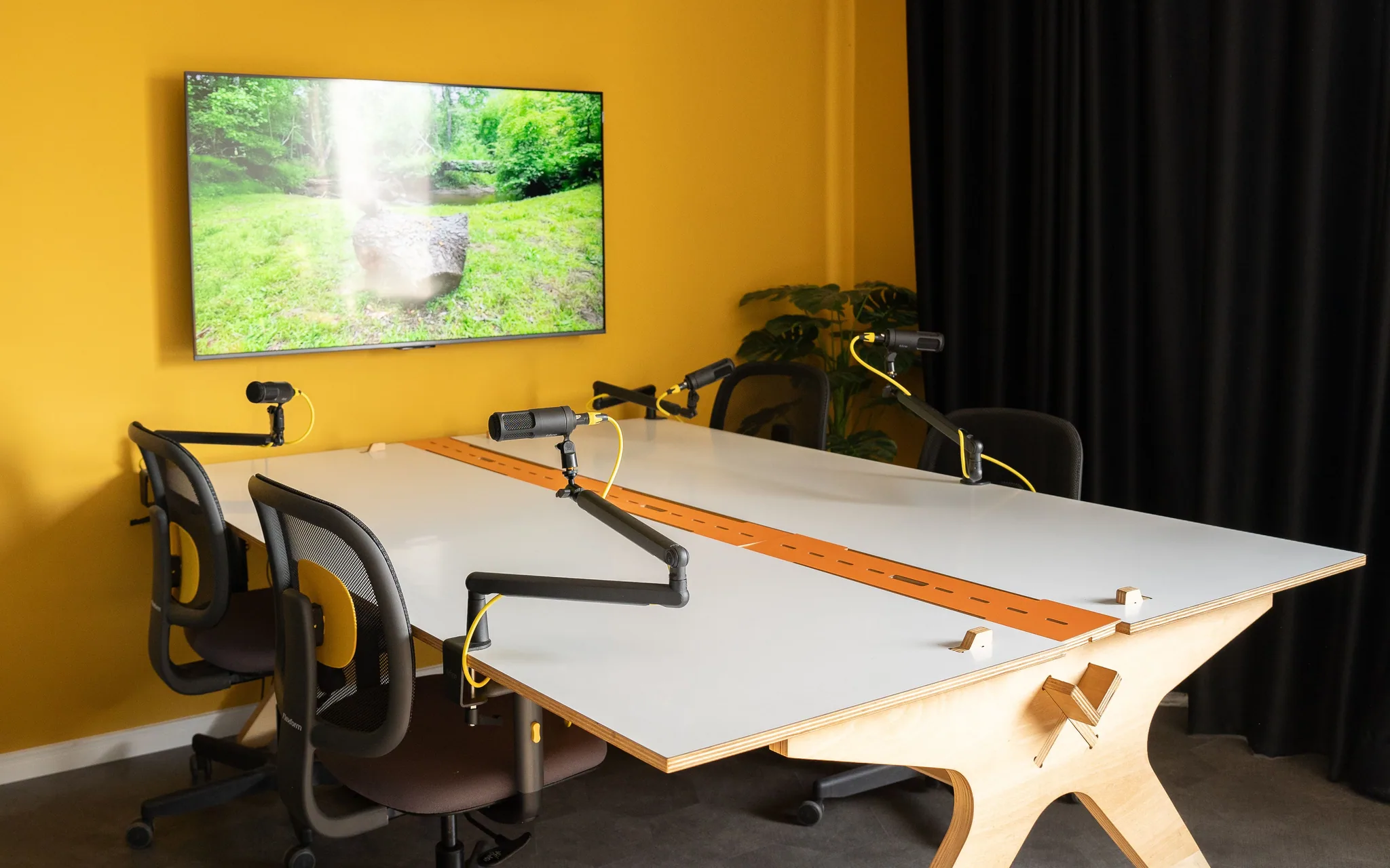 Mesa branca com friso laranja, quatro braços de microfone e quatro cadeiras, com a TV na parede amarela.