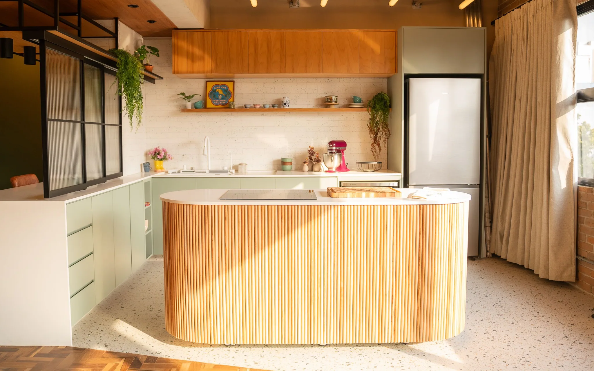 The whole kitchen with afternoon sun coming through the right-hand window, a beige curtain beside it and shadow drawn across the island.