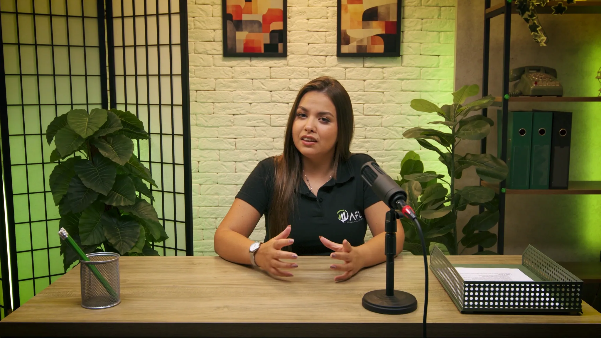 Cenário de curso em formato de entrevista: mesa de madeira, microfone de braço, biombo e plantas contra a parede de tijolo branco.
