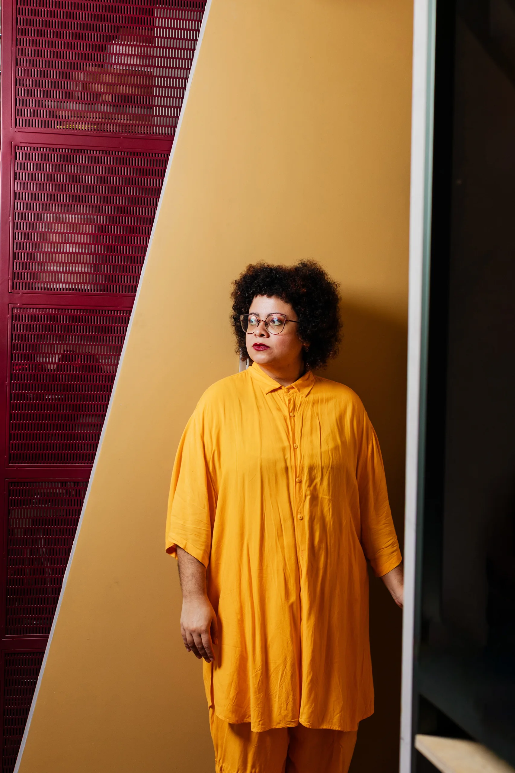Retrato contra parede mostarda ao lado de um locker vermelho, no Espaço Híbrido.
