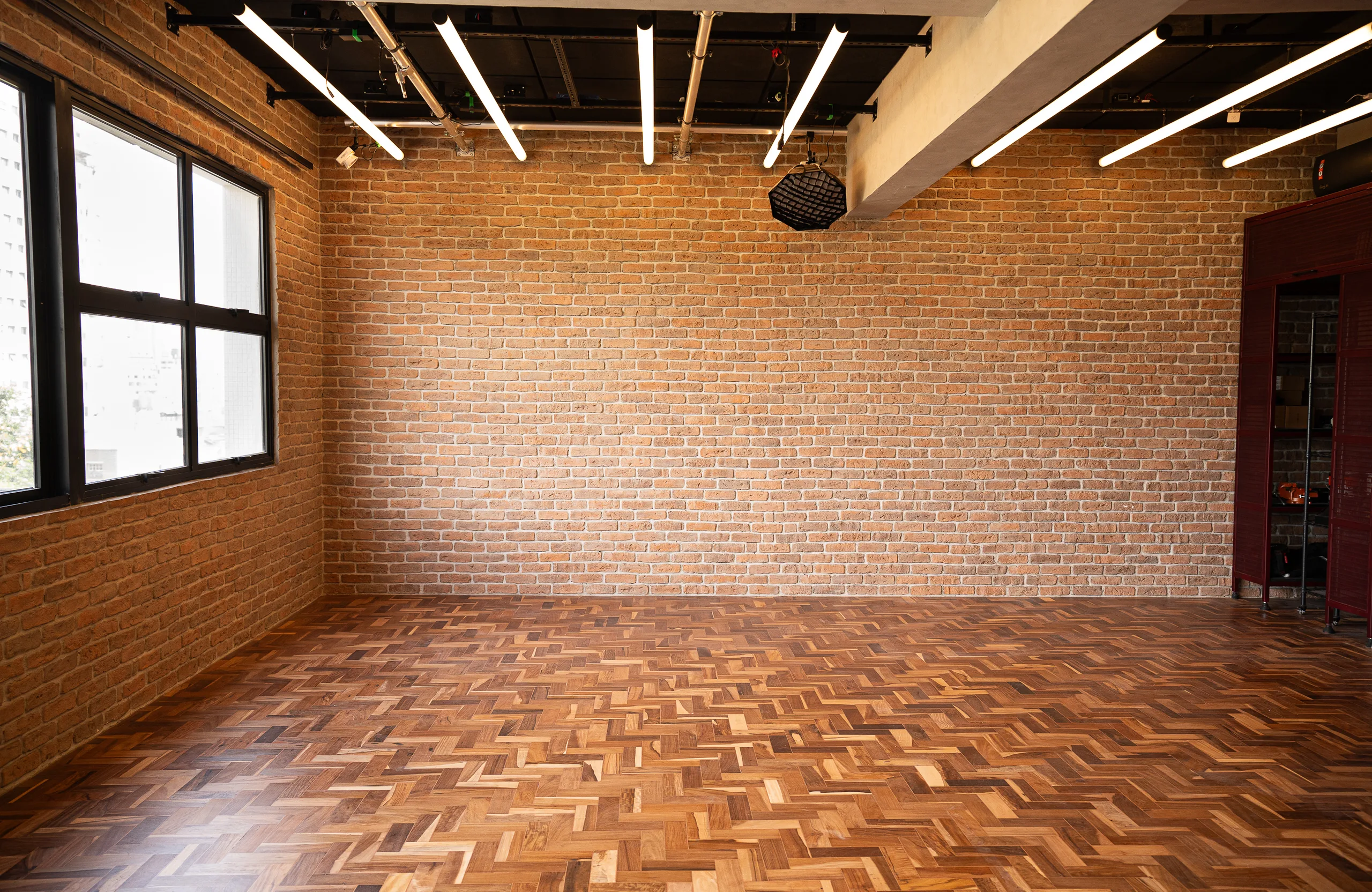 O Loft vazio: parede de tijolo aparente, taco espinha-de-peixe, janela ampla e trilhos de luz no teto.
